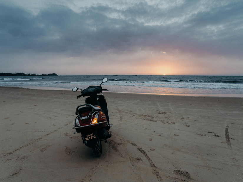 A scooter parked on a sandy beach at sunset with ocean waves, cloudy skies, and soft evening light in the background.