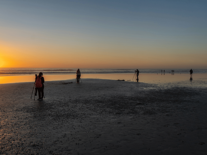 Distant human figures silhouetted against a fading golden horizon on a wide, reflective sandy beach during twilight.