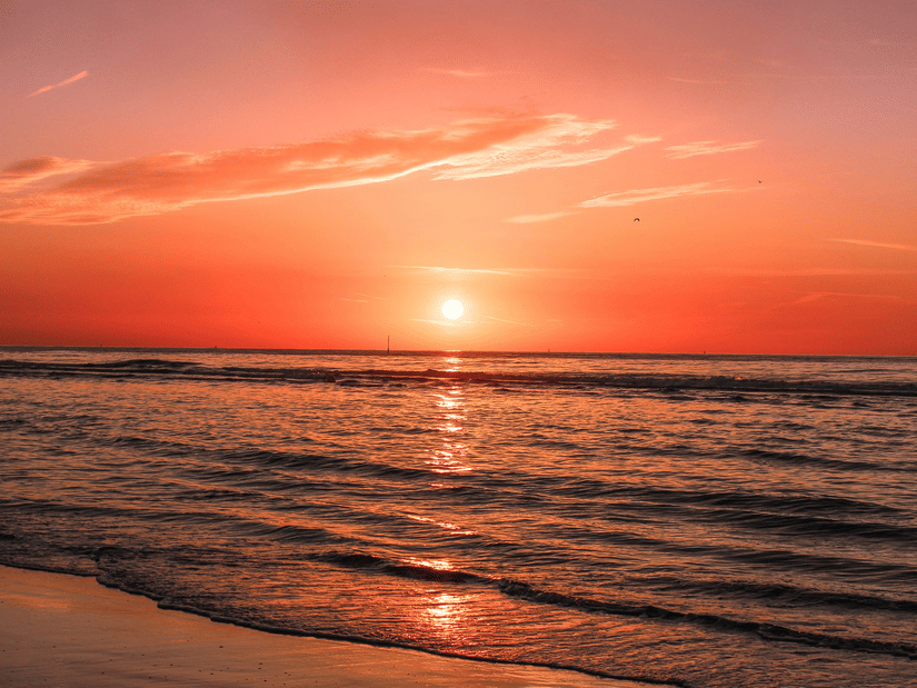 A serene orange sunset over a calm ocean, with gentle waves lapping onto the wet sand along the shoreline.