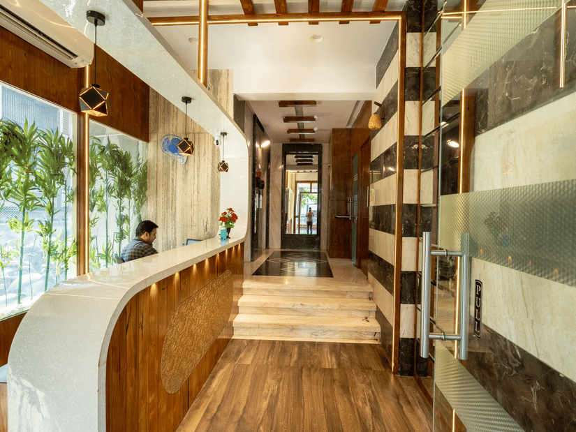 A reception area at Home2 India with a curved front desk, chairs, wall decor, large glass windows, wooden flooring, and a corridor leading inside.
