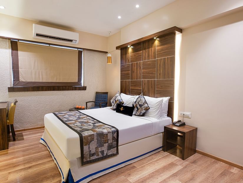 A side corner view of the Deluxe room at Hotel Bawa Regency, Dadar, featuring a plush bed, a window, a small stool beside the bed, and a working desk.