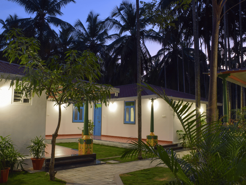 A white resort cottage with a red roof and blue window frames, surrounded by palm trees and green lawns - Ibex Resorts, Coimbatore (Kakarla)