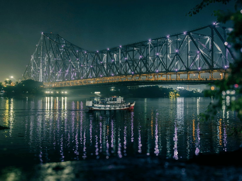 Howrah Bridge illuminated at night, reflecting on calm river waters, with boats drifting beneath and city lights shimmering softly