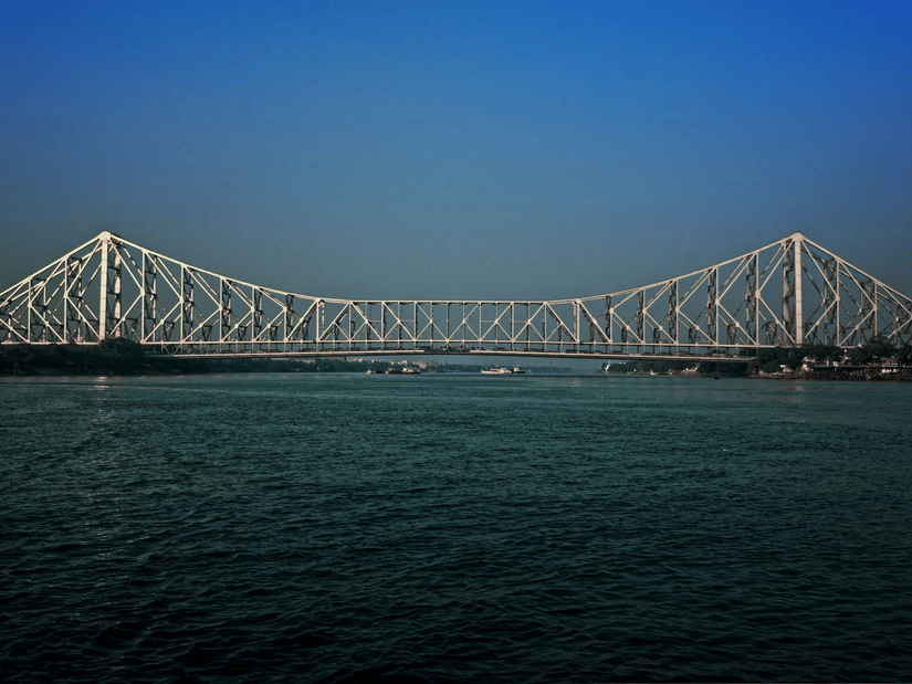 Howrah Bridge spanning the Hooghly River under a clear blue sky, with calm waters below and the steel structure stretching across