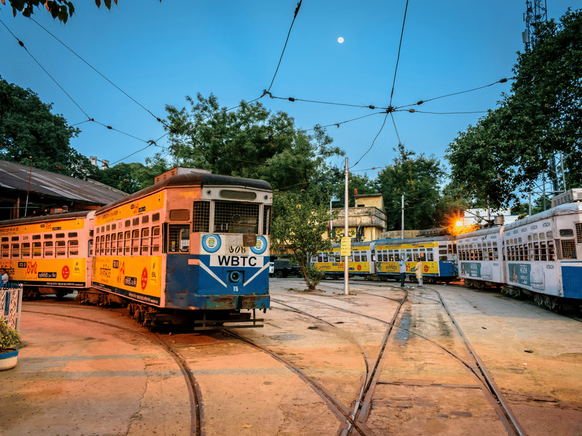 Two traditional Kolkata trams traveling side by side on parallel tracks through the city, showcasing the charm of Kolkata's iconic public transport system.