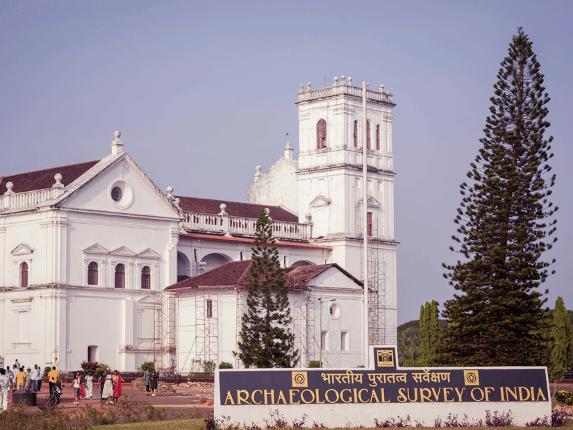A facade view of Se Cathedral Church in old Goa with many people walking on one side and a large Christmas tree on the other side, there is also a signage with the words Archaeological Survey of India in the foreground.
