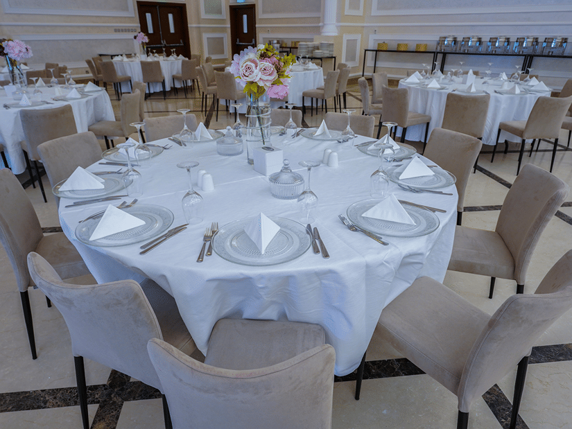 A banquet hall with round tables set for an event. Each table is covered with a white tablecloth and arranged with plates, napkins, and cutlery. Neutral-coloured chairs surround the tables, and a floral centrepiece adorns each at La Maison Hotel, Doha.
