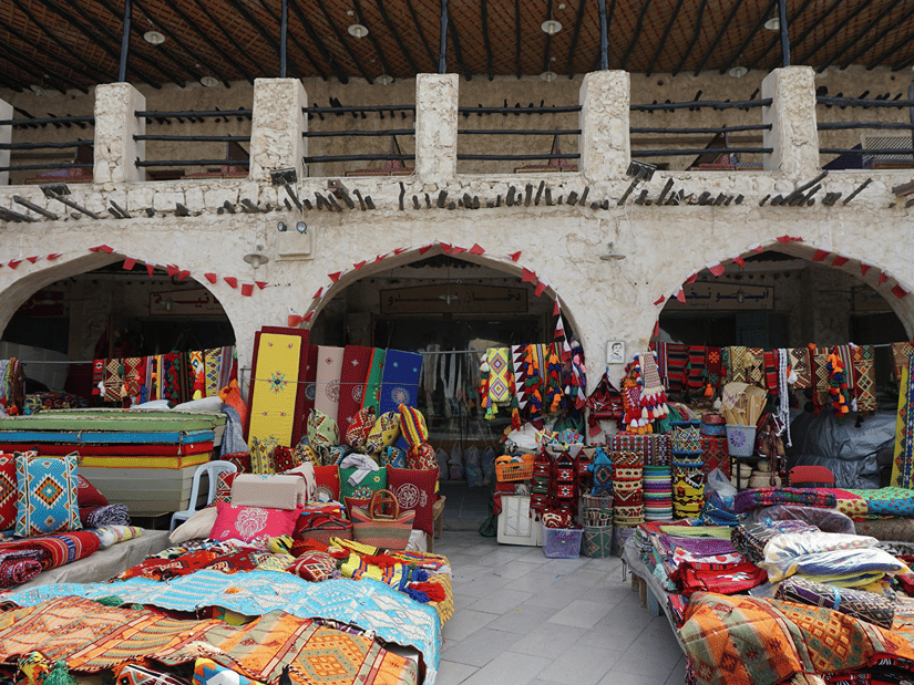 An overview of Souq Waqif market with people selling rugs in a courtyard and an old building in the background.