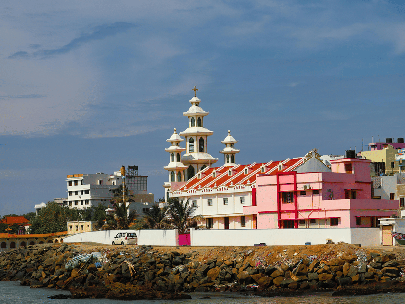 A view of a white and pink multi-tiered church building situated on a rocky coastline against a clear blue sky.