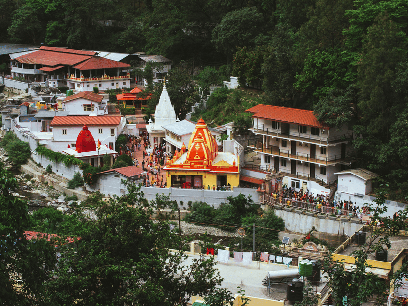 A scenic view of the colourful Kainchi Dham with red and white domes nestled in a lush green valley by a river.