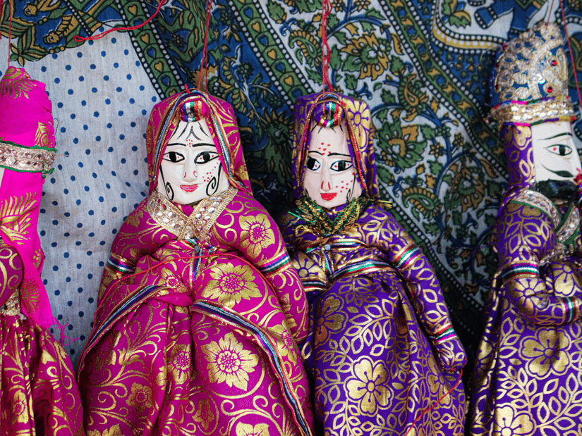 Kalbelia dancer twirls gracefully beside palm trees at sunset, colourful skirt spreading dramaticallyTraditional Rajasthani puppets dressed in ornate pink and purple costumes hang against patterned fabric