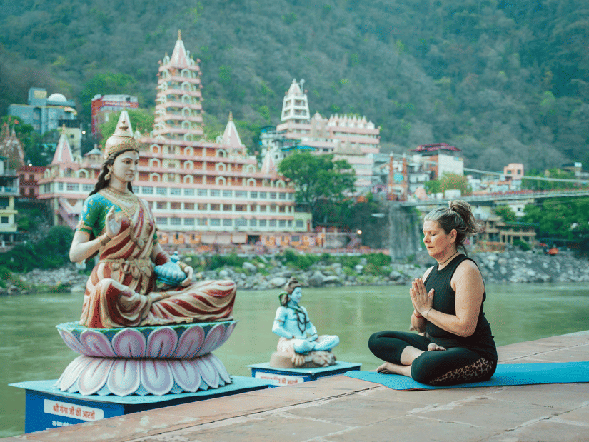 A woman practicing yoga in a seated pose by a river, with large Hindu statues and a multi-storey temple behind her.