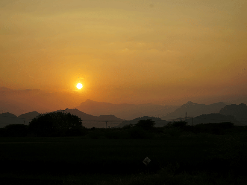 A wide landscape shot showing a bright sun setting or rising behind the silhouetted peaks of a mountain range under a warm, hazy sky.