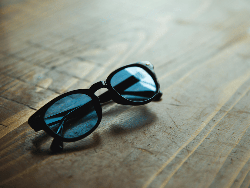 A pair of dark-framed sunglasses with blue-tinted lenses resting on a textured wooden surface.
