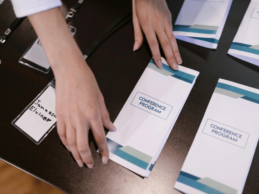 Close-up of hands arranging white and blue conference programme leaflets and ID badges on a dark tabletop.