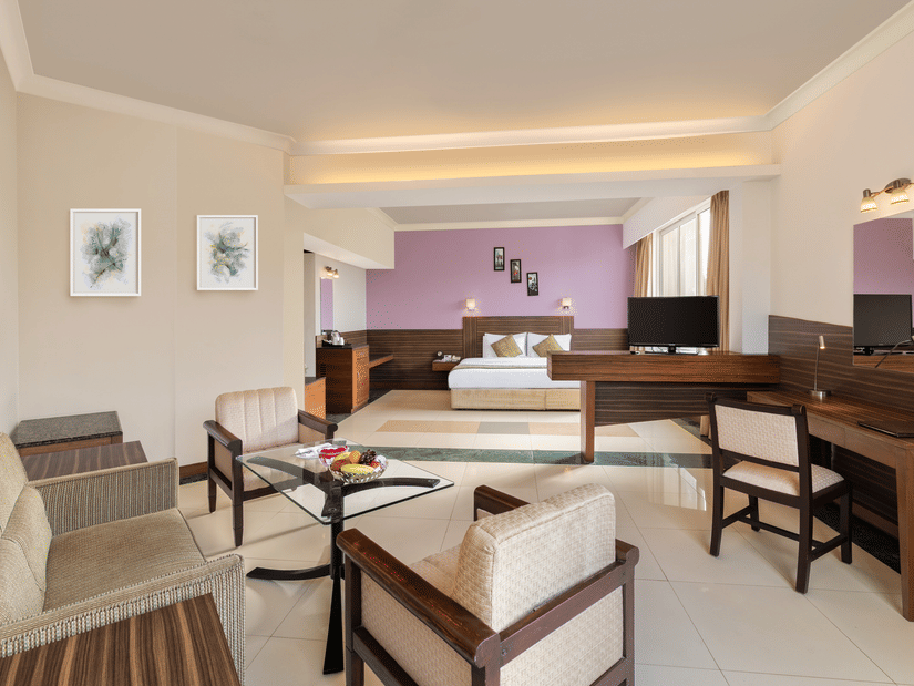 An overview of the Deluxe Rooms in Tanjore at Sangam Hotel, Thanjavur, featuring a double bed in the background with lamps, paintings, and a study table, chairs, and a coffee table in the foreground.