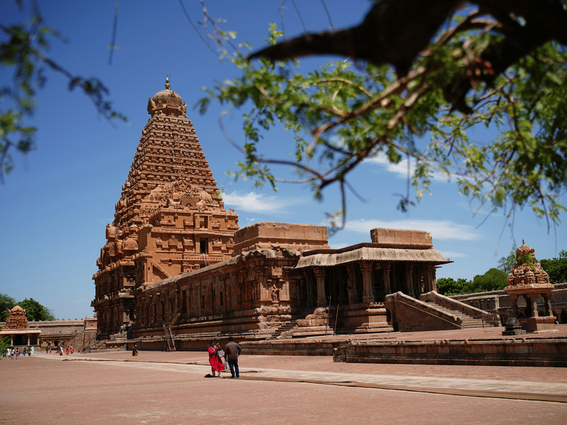 The grand Brihadeeswarar Temple complex stands prominently under a clear blue sky with an open space next to the temple and people walking on it. This temple is one of the popular places to visit during a Thanjavur trip from Chennai.