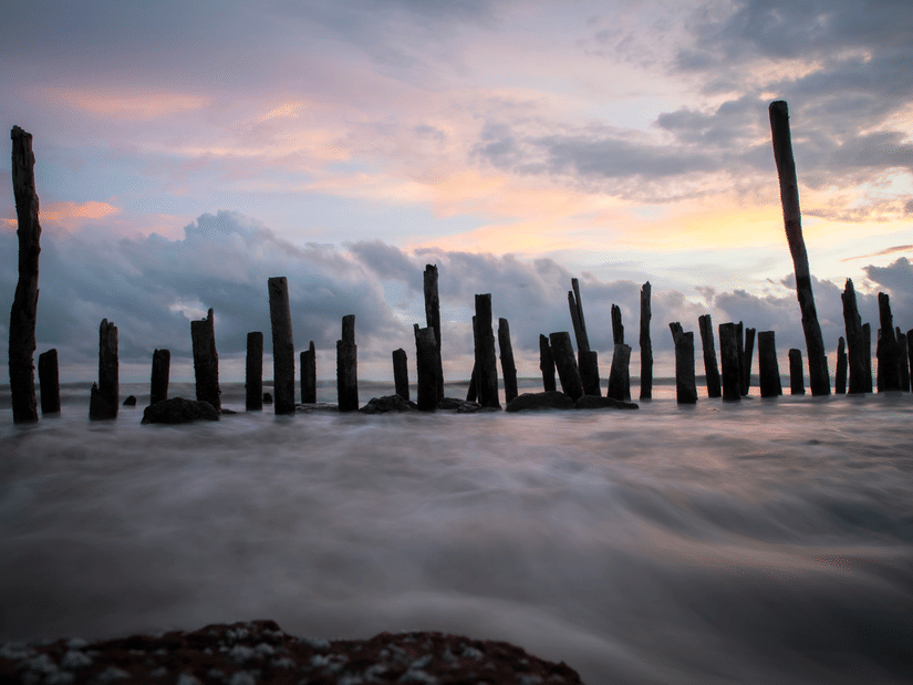 Weathered wooden posts stand tall along the sea shoreline of Tajpur Beach as gentle ocean waves wash past under a colourful sunset sky.
