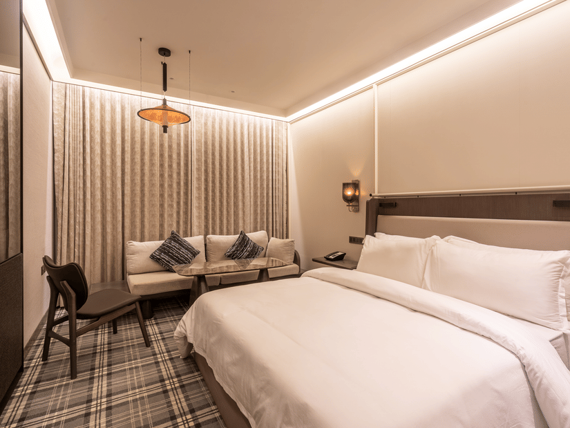 Modern hotel room at The Residency Towers Chennai featuring a large bed, seating area with sofa and chair, patterned carpet, and warm ambient lighting.