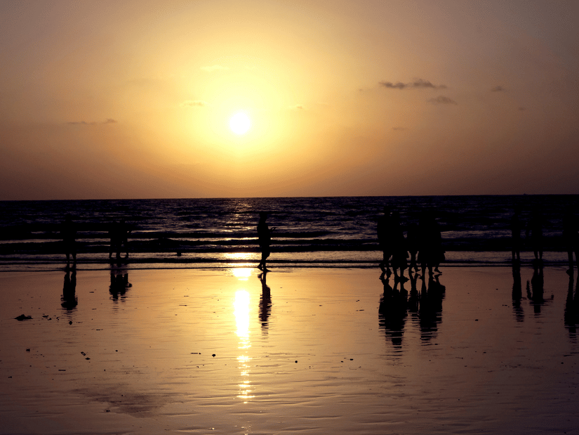 A view from the shoreline of the Aksa Beach sunset, with numerous people standing on the shore and waves overlapping it