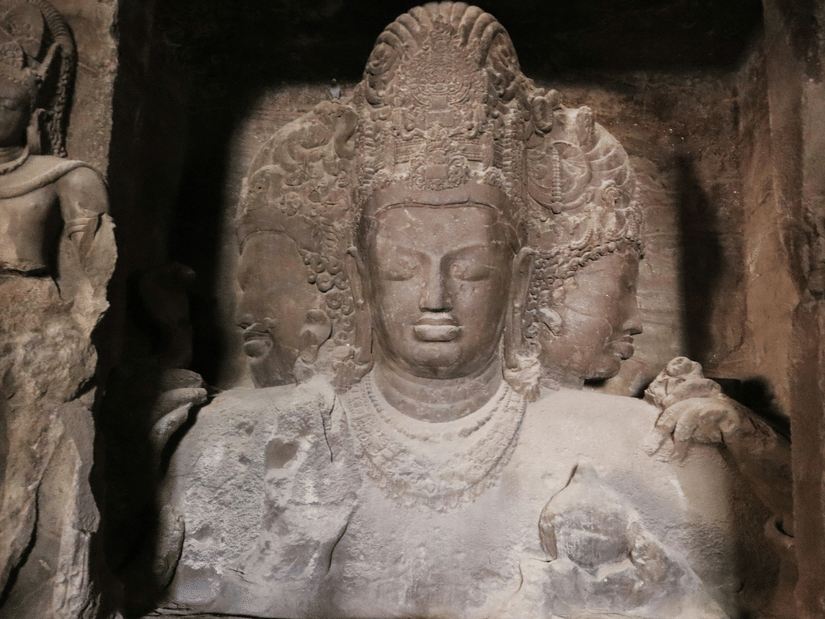 Multi-faced stone sculpture of a deity inside Elephanta Caves, with intricate carvings and weathered rock surfaces, captured during a day tour in Mumbai.