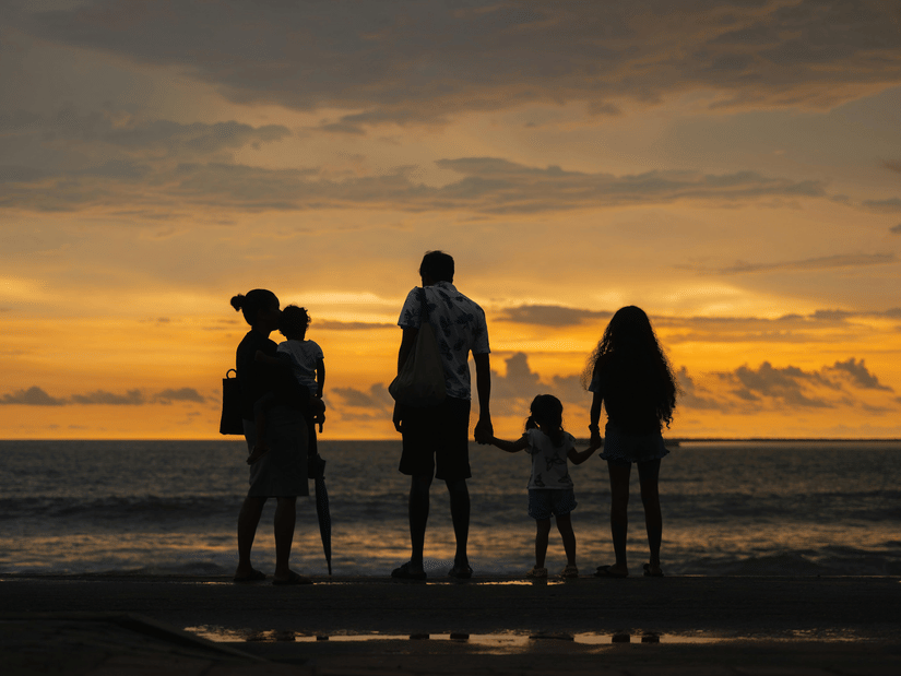 Family of five holding hands on the shore at sunset with dramatic orange and yellow sky reflecting on the water