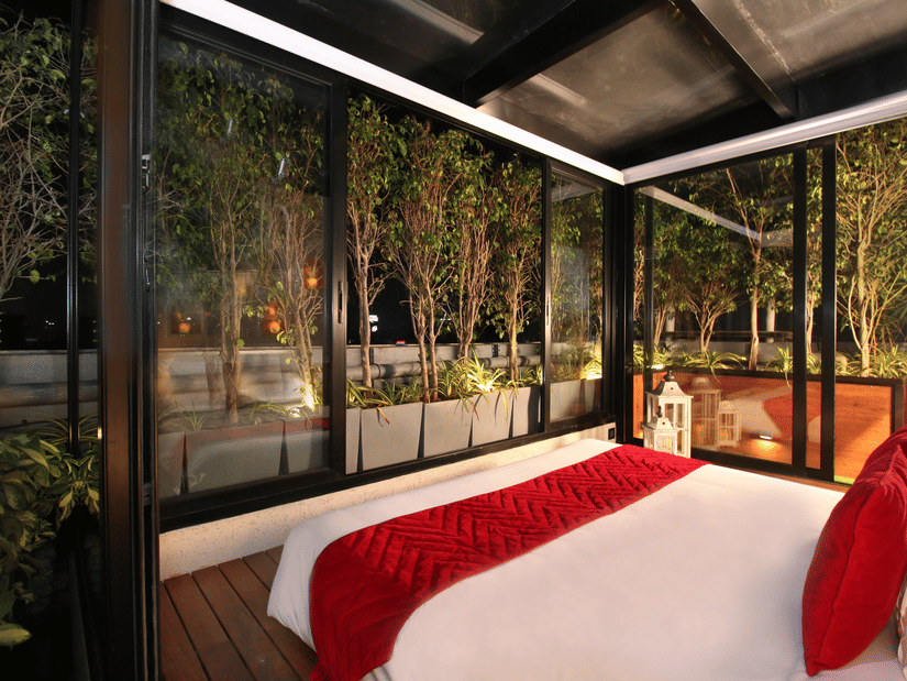 A bedroom at Theory9 - Premium Service Apartments, Bandra with a large bed and floor-to-ceiling windows overlooking a night-time rooftop garden with trees