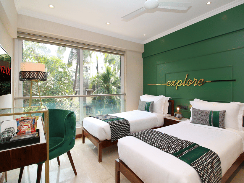 Twin beds facing the TV on the wall with a table and chair below, and large windows by the bed at Theory9 - Premium Srervice Apartments, Bandra