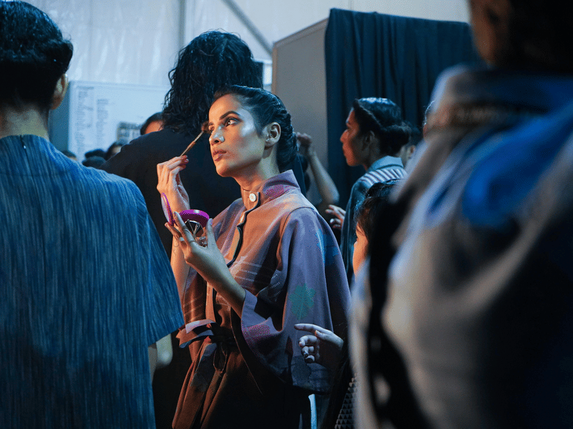 A model applies makeup backstage at a fashion show, surrounded by a busy crowd in a dimly lit, cool-toned room.