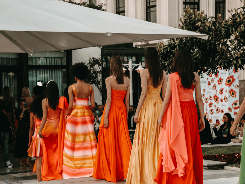 Models in vibrant orange and yellow summer dresses walk an outdoor catwalk beneath a large white canopy at a fashion event.