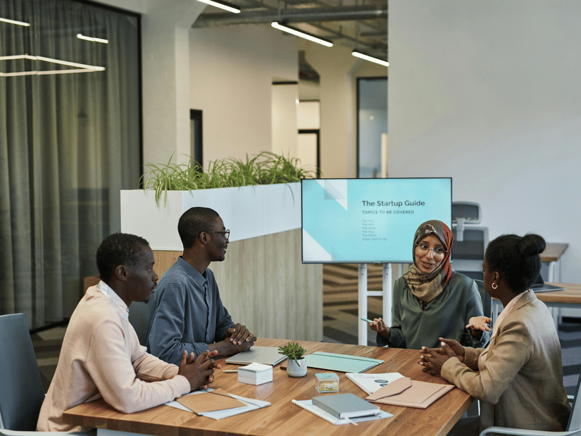 Four professionals having a meeting around a table, discussing 'The Startup Guide'displayed on a screen, with notebooks and a small plant on the table.