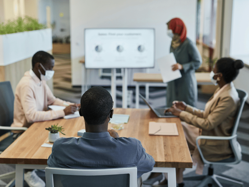 Four coworkers wearing face masks sit around a table during a meeting while a colleague stands and presents data on a screen in a modern office.