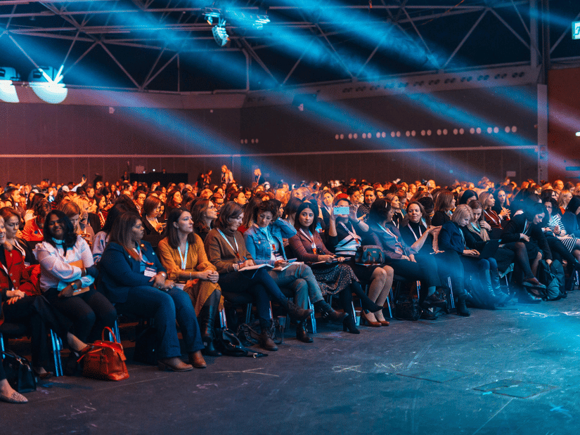 A view from the podium of the numerous people sitting on chairs and listening to a conference with spotlight rays shining down.