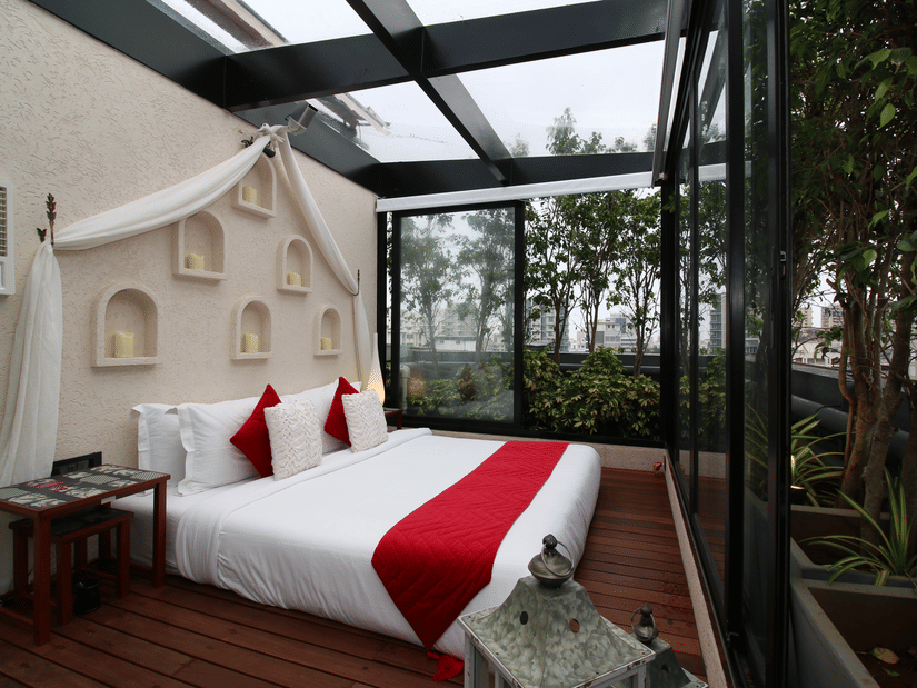 An overview of the Night Under the Sky Room with glass walls and ceilings, a double bed, side tables and AC with trees in the background at Theory9 - Premium Serviced Apartments, Bandra.