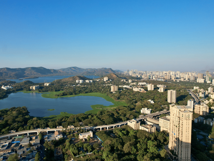 An aerial view of Powai Lake with hills in the distance and a metro line and apartment complex in the foreground.