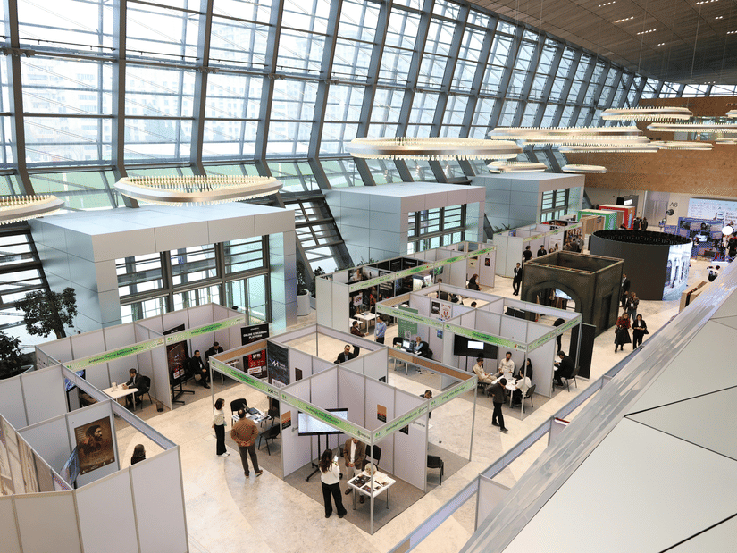 An overview of a trade show with stalls aligned next to each other and people walking around looking at things.