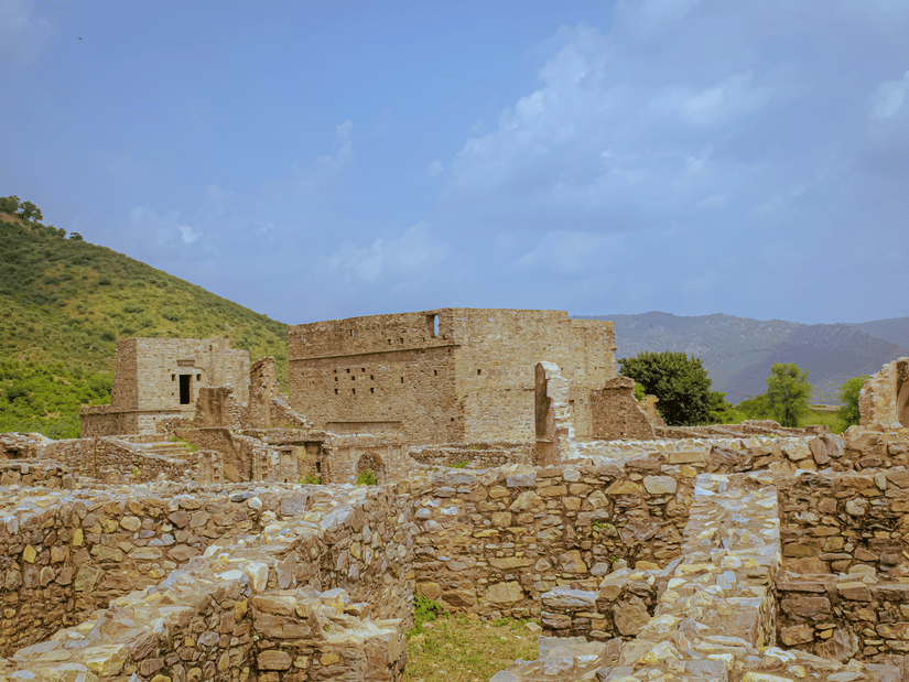A view from inside Bhangarh Fort of the remains of the stone houses, a mountain on the left hand side and the blue sky in the background.
