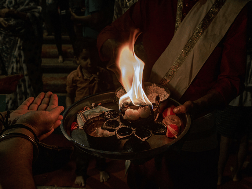 A priest’s hands hold a lit ceremonial flame during a Hindu worship ritual, with the fire illuminating the surrounding scene.