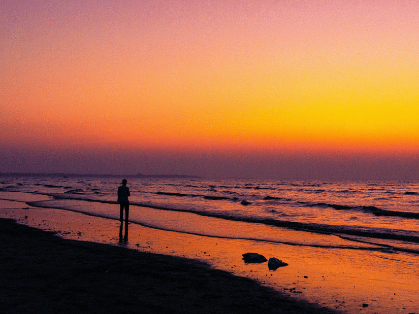 Vibrant sunset over Daman Beach with gentle waves, colourful skies, and a visitor standing along the water's edge