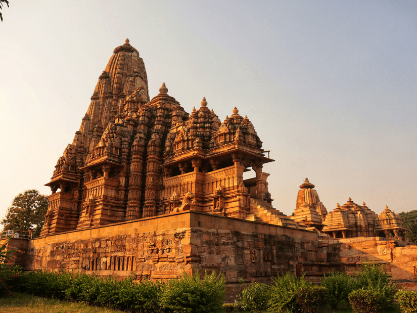 A view of a Indian temple with intricate art on the exterior wall, surrounded by greenery, as seen during day time, symbolising Shree Bhrigu Rishi Temple.