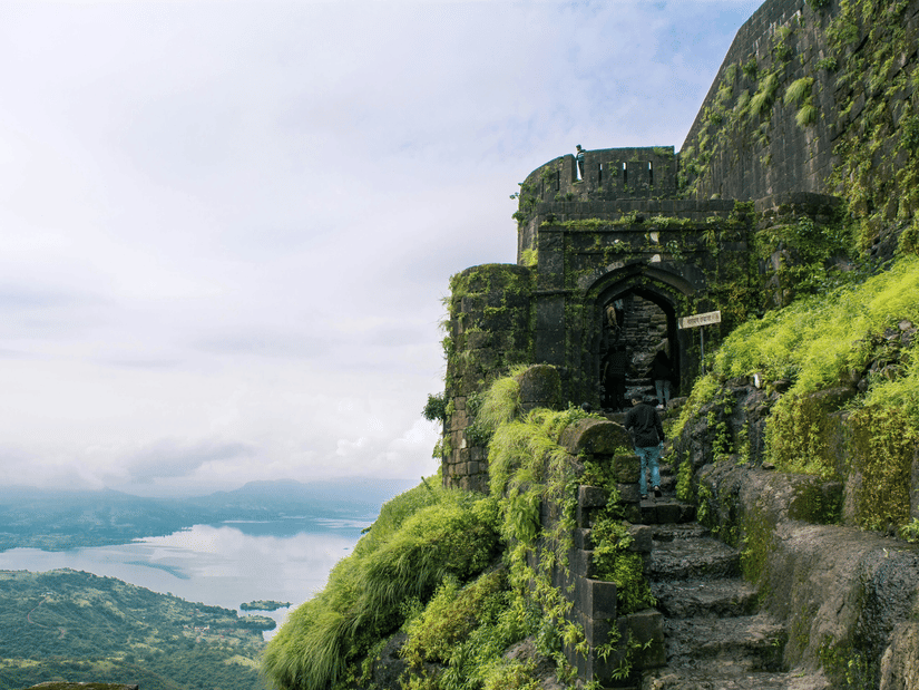 A view from afar of the steps covered by moss and grass on the way to Lohagadh Fort in Lonavala with a lake and clouds seen in the distance.