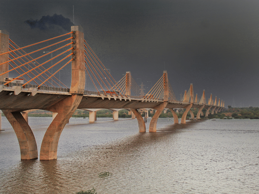 A view of the Narmada Cable Bridge, one of the best places to visit in Bharuch, over the Narmada River with a golden hue on it and dark clouds above