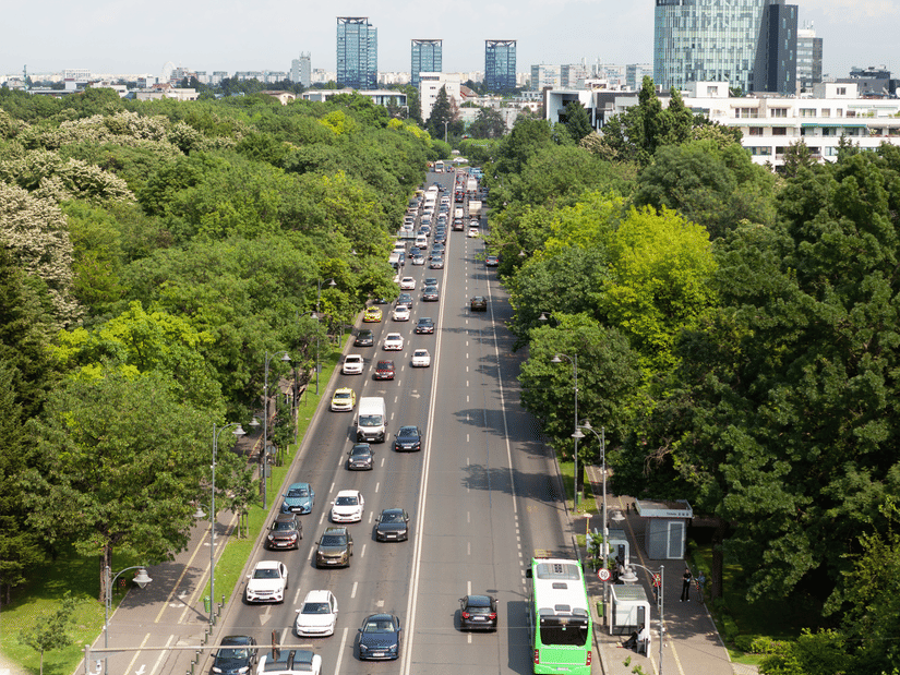 An elevated view of a multi-lane city road with vehicular traffic, lined by dense green trees on both sides with high-rise buildings in the distance.