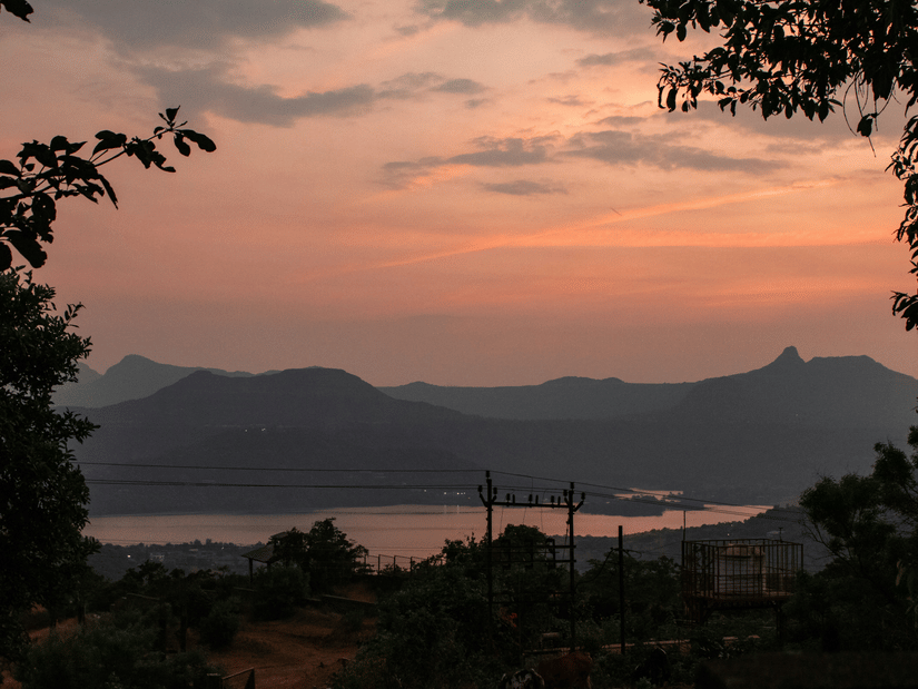 An overview of Pawna Lake in Lonavala, as seen during twilight hour with orangish hues in the sky and mountains in the background. This camping site is one of the ideal Lonavala things to do at night with bonfires.