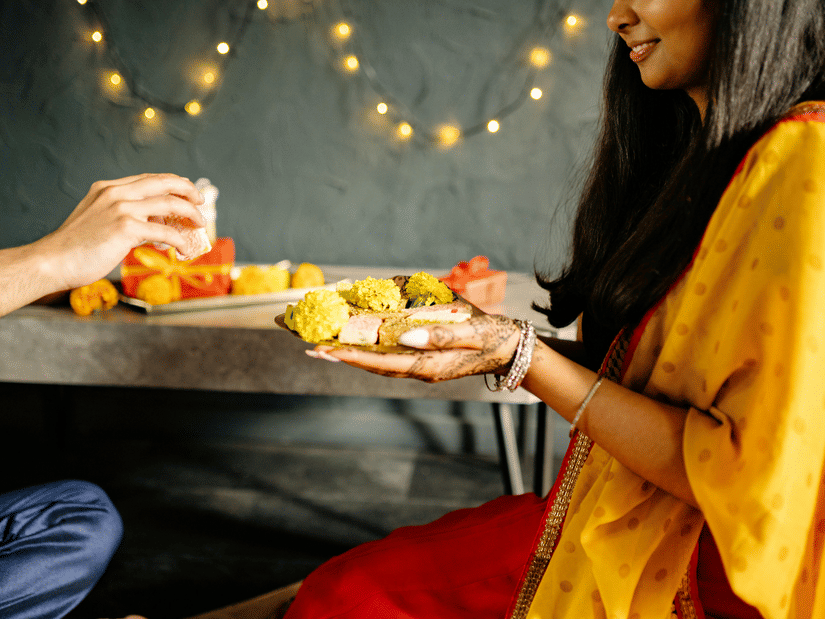 2 people in traditional attire are seated at a table, sharing colourful Indian sweets.
