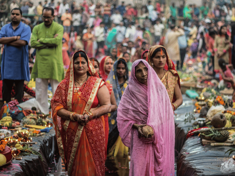 A crowd of people participating in a cultural or religious festival; women in colourful traditional saris stand in the foreground near offerings.