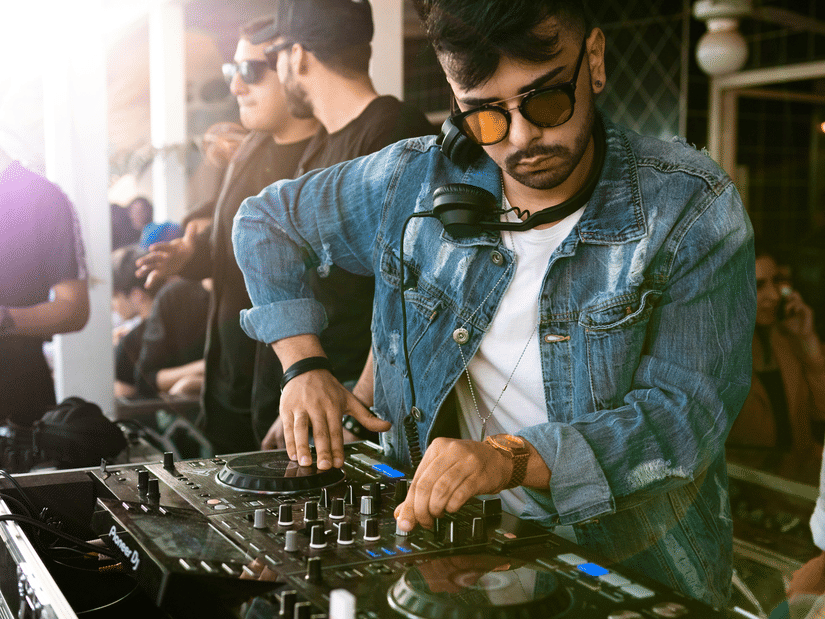 A man with sunglasses and a denim jacket operating a DJ mixer setup surrounded by people.