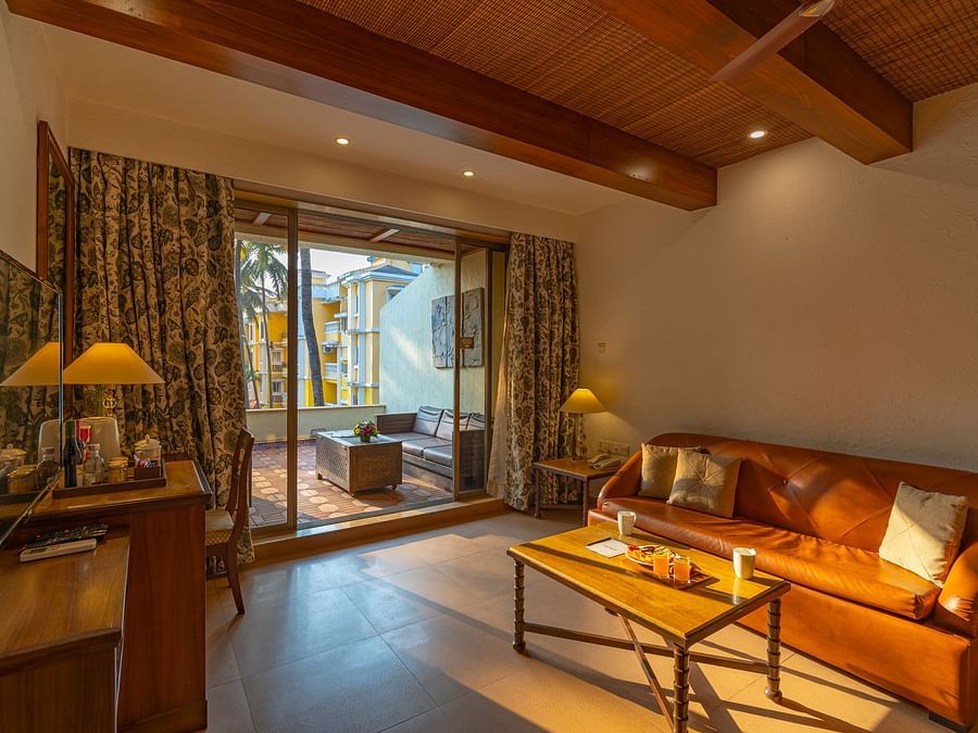 A living room that includes a sofa, coffee table, tea and coffee maker, a TV, and an outdoor balcony with seating arrangements.