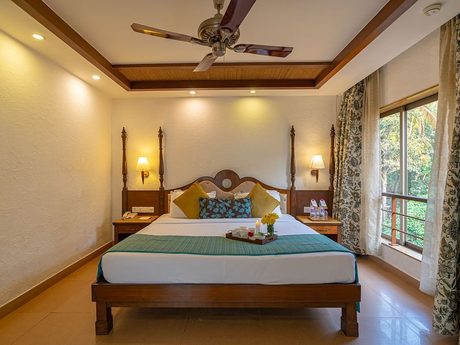 An image of a hotel room featuring a beautifully decorated bed with a tray of cups, plates, and flowers, complemented by a large window, a fan, and table lamps.