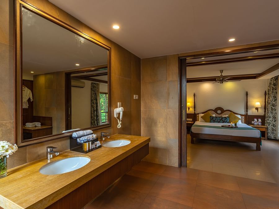 An image of a washroom featuring a large square mirror, a sink, towels, and shower amenities, with an open door revealing a room that includes a bed.